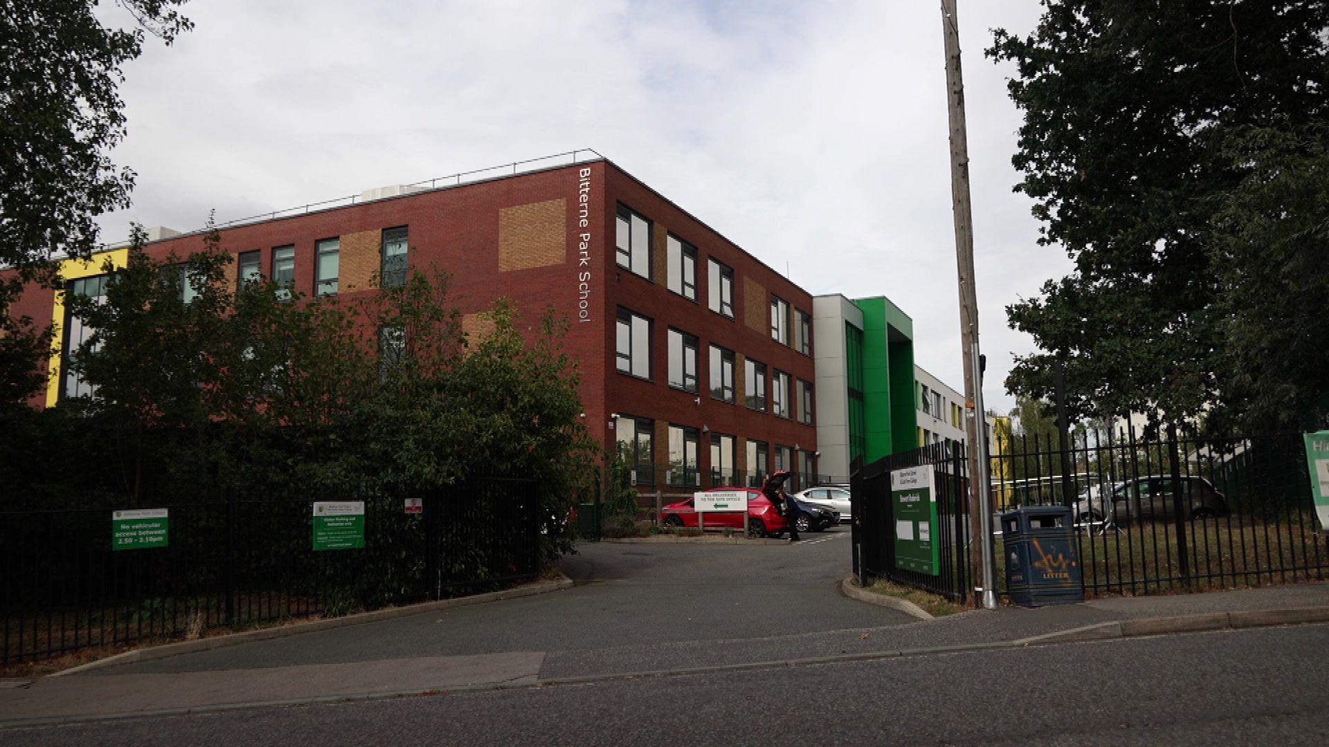 An exterior view of Bitterne Park School. It is a three storey brick building with a green entrance and the schools name in a sign on the side. It is surrounded by a black fence.
