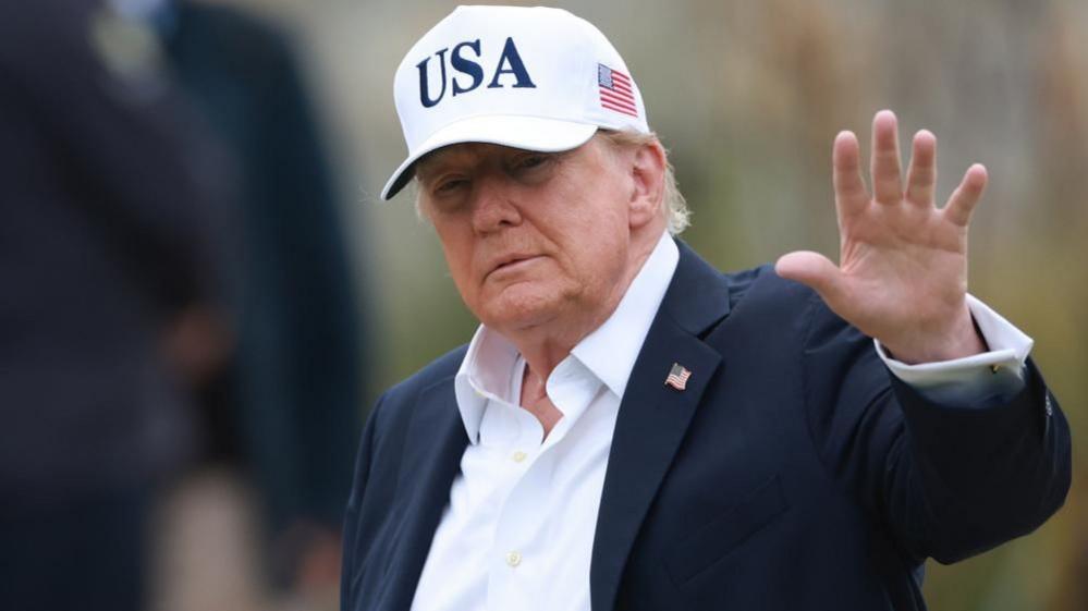 Donald Trump is looking at the camera. He is wearing a white baseball cap with the letters USA emblazoned on the front in blue.