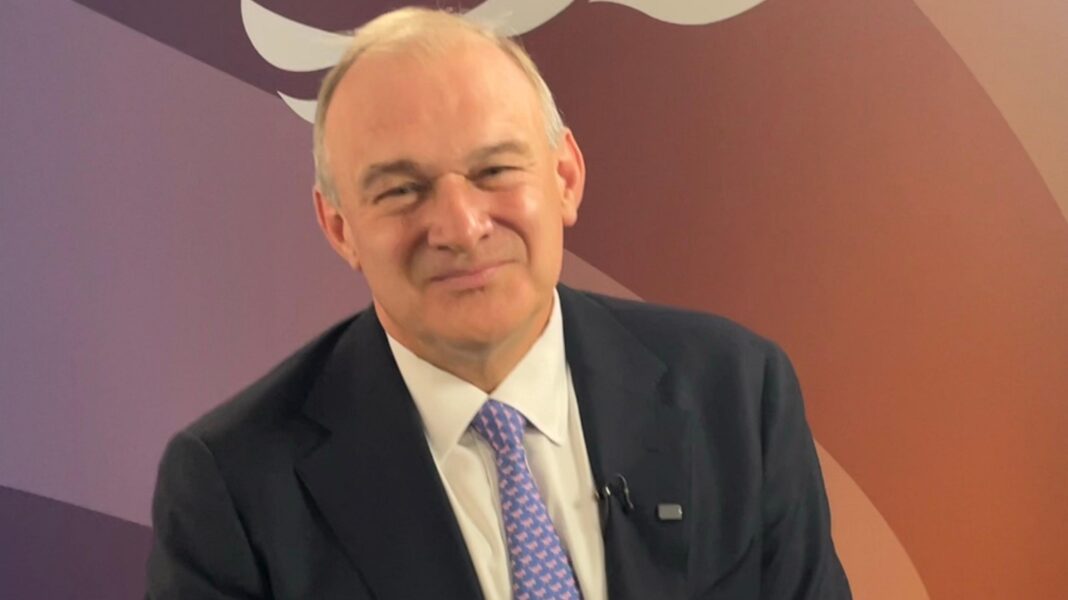 Brexit to blame for small boat migrants, says Lib Dem leader Sir Ed Davey, an older man wearing a dark suit, white shirt and patterned lilac tie. He is sat in front of the Liberal Democrats logo, depicting a bird.