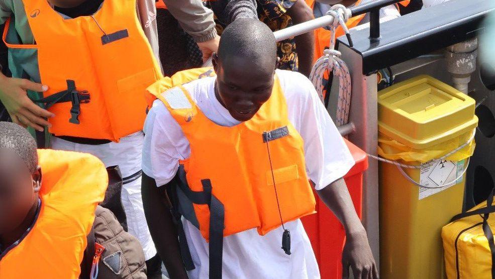 A man with a shaved head coming off a boat wearing a white shirt under an orange life jacket.