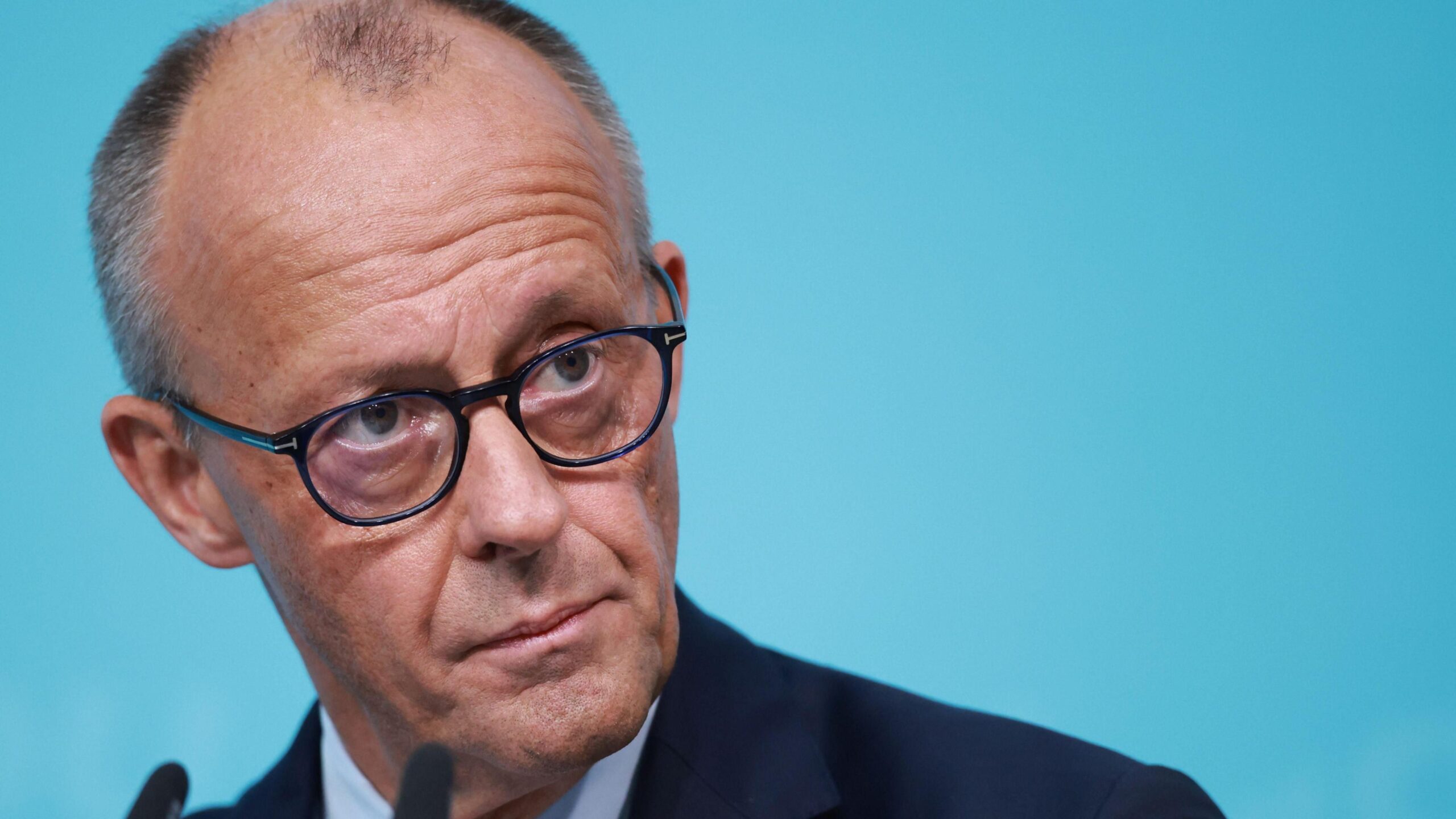 Friedrich Merz pictured on stage. He has short, grey hair and wears round, black-framed glasses. He is stood in front of a bright blue background and is leaning towards two small microphones. He is looking towards the right side of the frame.