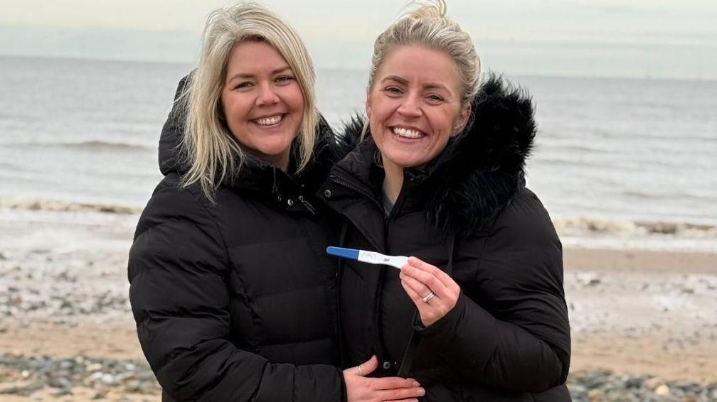 Two blonde, white women, one with her hair tied up and the other with her hair down, in big black coats, stand side by side with their arms around each other. Both are smiling and one of them is holding up a pregnancy test and her wedding ring is visible. They are stood on a beach and in the background there is sand, rocks, as well as the sea.