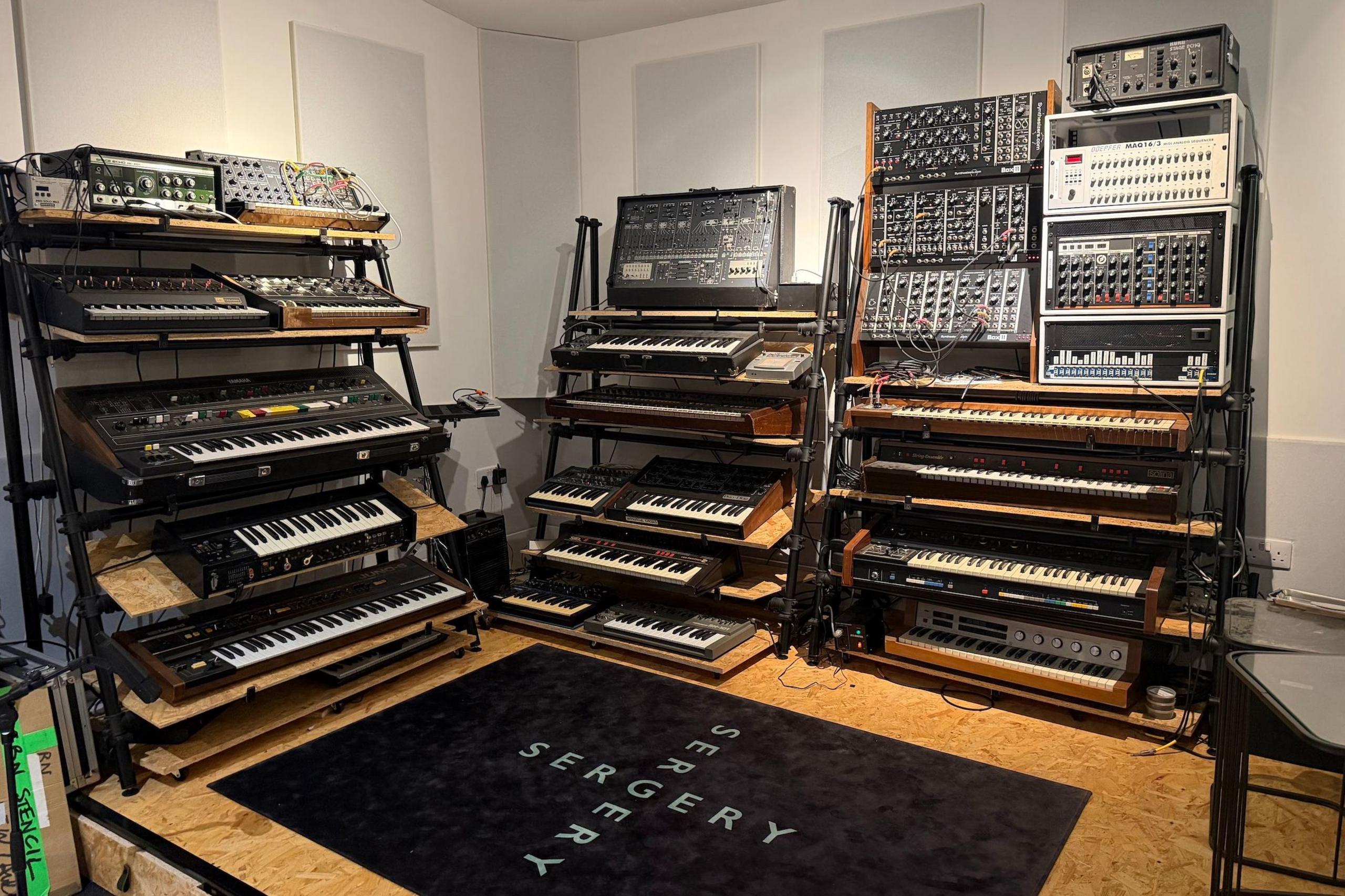 Two walls of synths, inside Serge Pizzorno's home studio. On the floor in front of them is a rug showing the studio's logo, "Sergery".