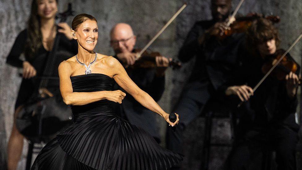 Dion dancing in a black dress, holding a microphone, while musicians play behind her