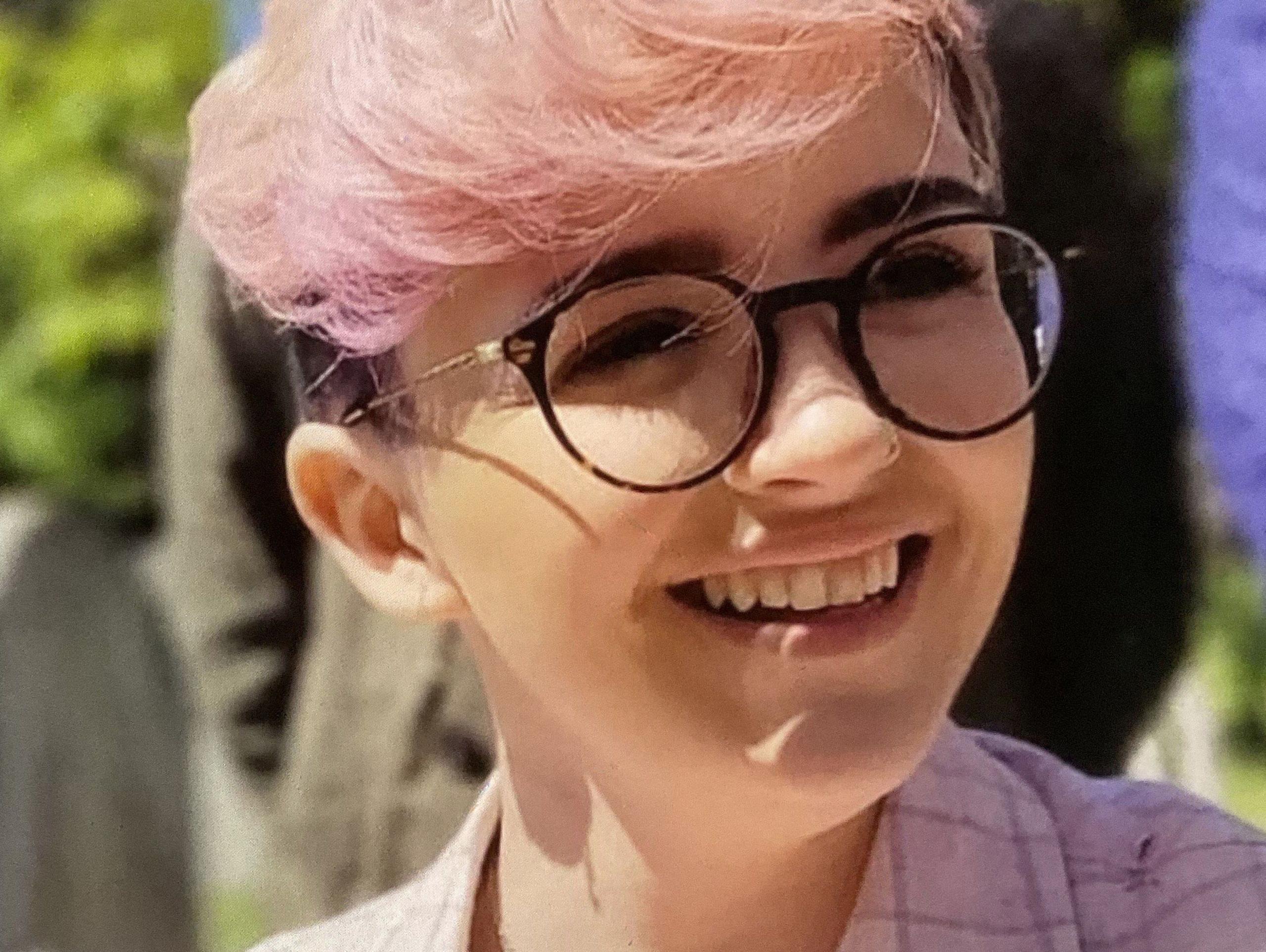 Imogen Nunn is smiling and looking off camera. She has pale pink hair and a pink checked jacket on, and is wearing dark rimmed round glasses.