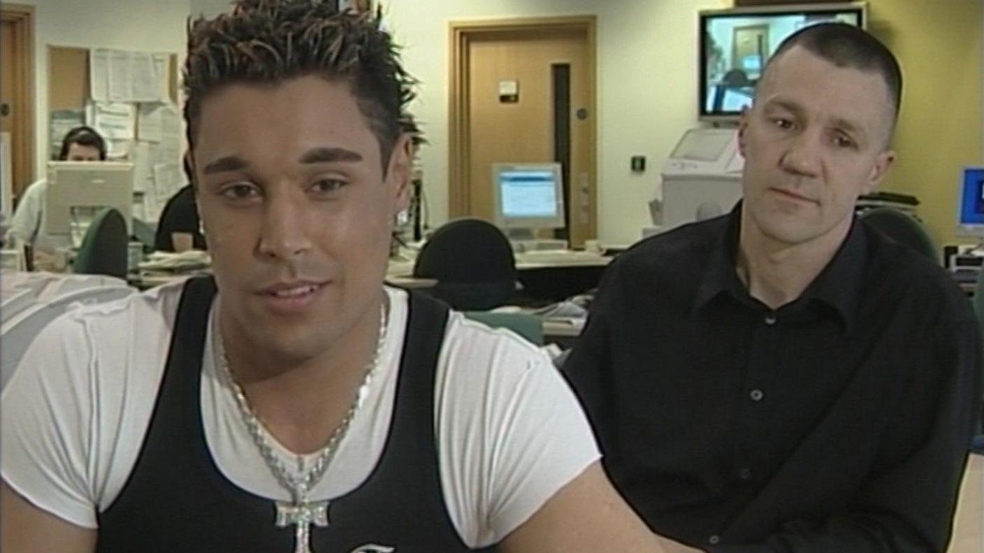 A man with spiky black hair with blonde tints and a silver chain looks into the camera. Another man with short hair wearing a black shirt looks down.