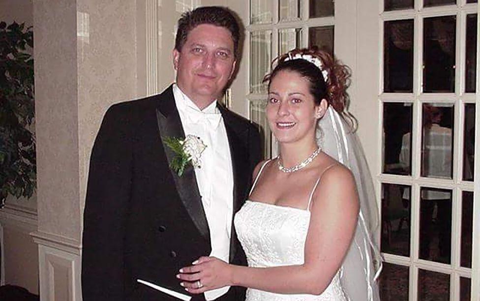 John on the left is in his wedding suit with a flower in his lapel. Sylvia is on the right in a wedding dress and tiara. They are both smiling at the camera.