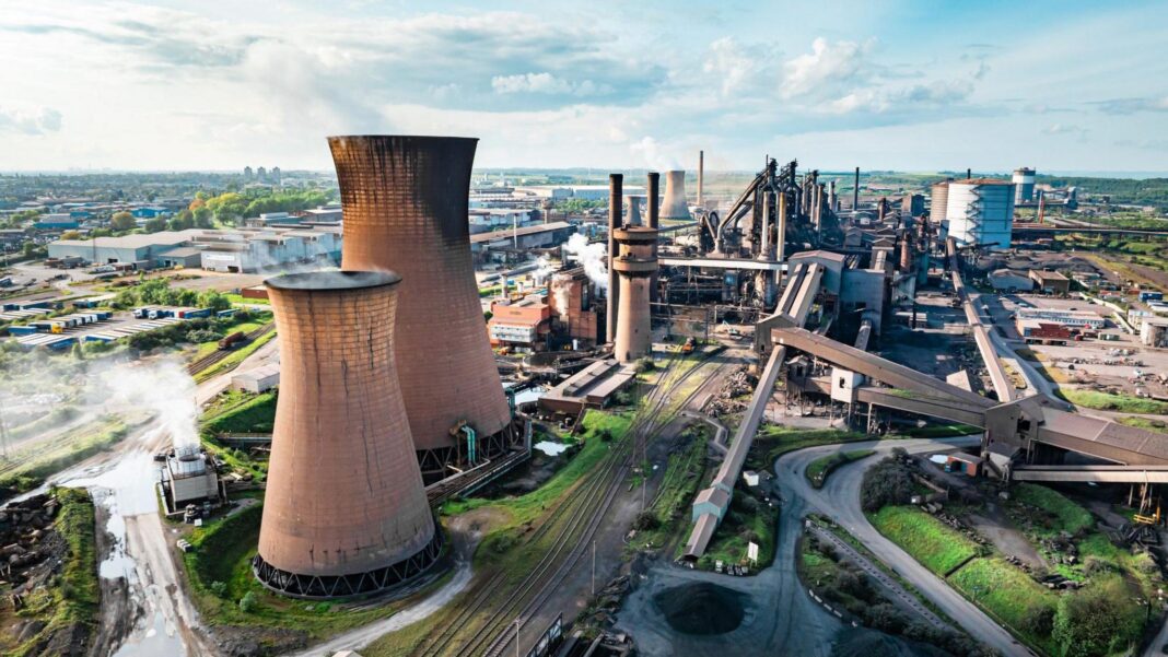 British Steel plant at Scunthorpe with towering cooling towers and smoke-stacks emitting steam. Conveyor belts and railway tracks weave through the industrial landscape. There are warehouses, roads and patches of grass. The sky is bright with scattered clouds.