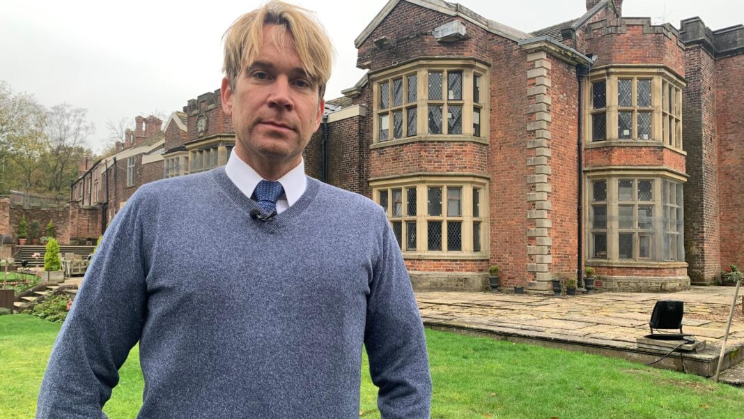 Hopwood DePree stands outside Hopwood Hall, in Middleton, Rochdale. He has blond hair and is wearing a tie under a blue jumper.