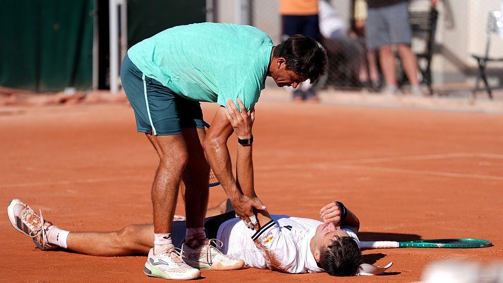 Jakub Mensik lays on the clay at Roland Garros after suffering in the heat during his 2026 French Open five-set victory over Mario Navone, who stands over him