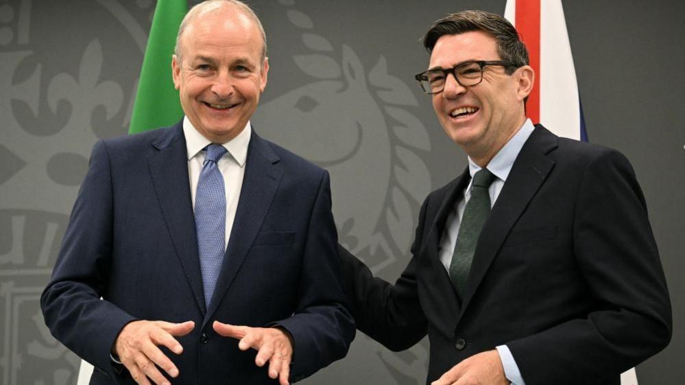 Martin is laughing on the left and Burnham is laughing on the right. Martin is wearing a navy suit and blue tie, he is bald. Burnham is wearing a black suit and green tie, he has black hair and wears glasses.