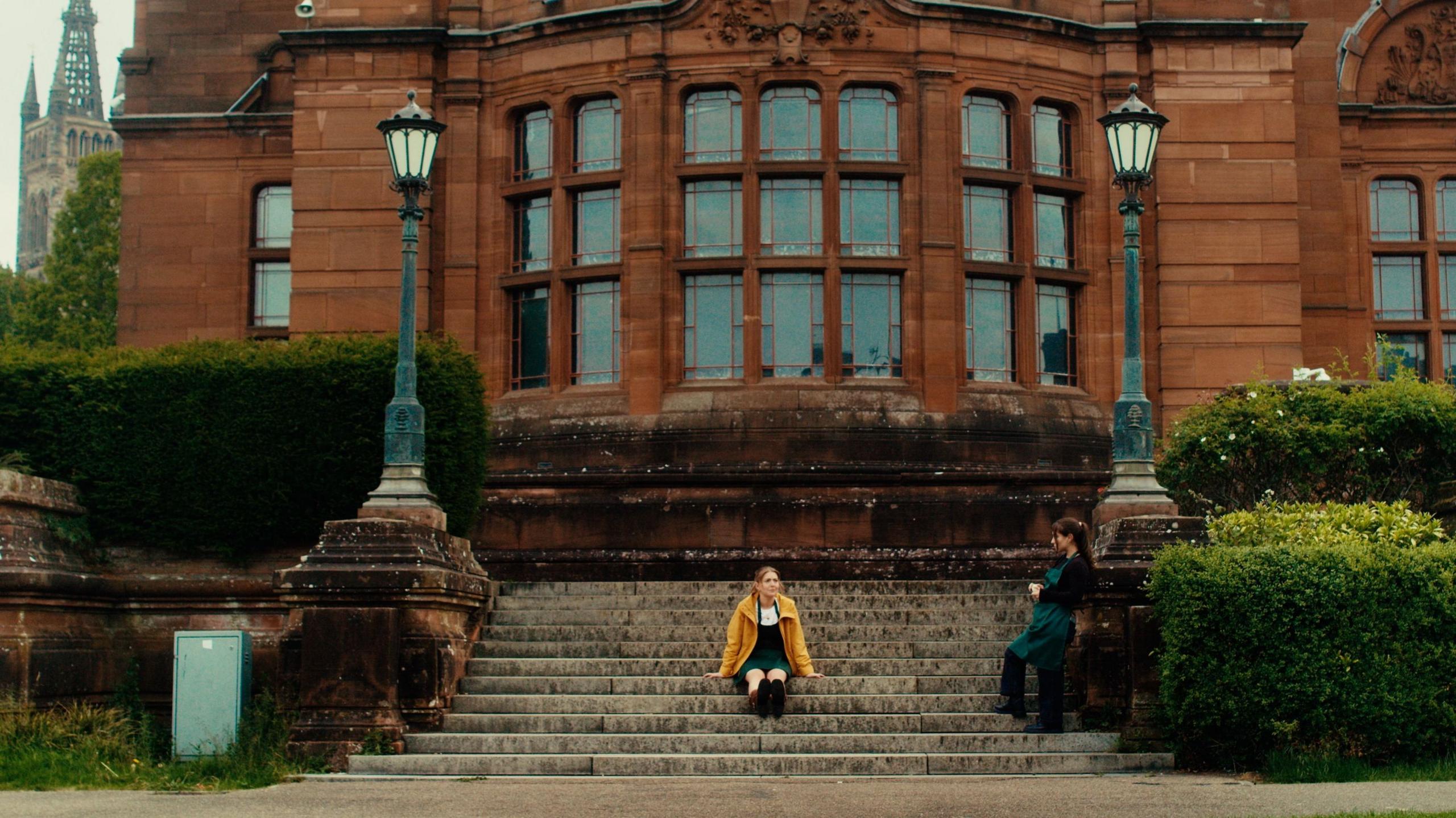 A wide view of a grand red sandstone building - the Kelvingrove Art Gallery - with large bay windows and ornate architectural details. Broad stone steps lead up to the entrance, framed by decorative lamp posts and neatly trimmed hedges. A woman sits on the steps wearing a bright yellow coat, while another woman stands nearby