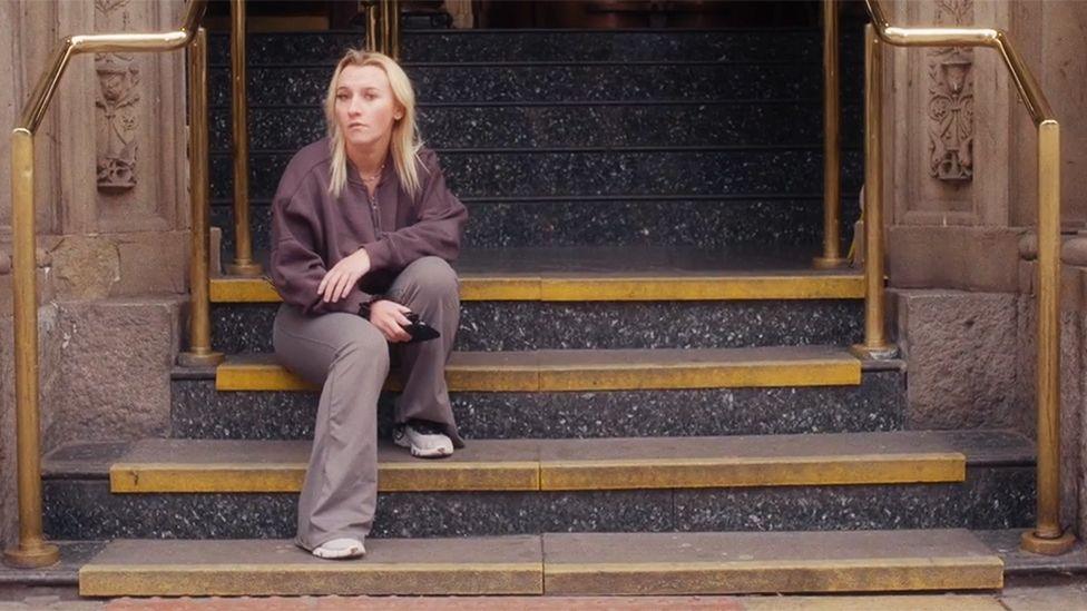 Holly, in casual clothing, sits alone on wide stone steps outside a building entrance. Brass handrails frame the staircase, and dark polished steps lead up to large doors behind her. Holly is holding a phone and looking away from the camera, with the architectural details of the entrance visible in the background.