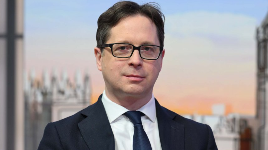 Head and shoulders shot of Burghart on the Sunday with Laura Kuenssberg set. He is wearing a black suit with a white shirt and a shiny navy tie. He has floppy brown hair and wears spectacles.