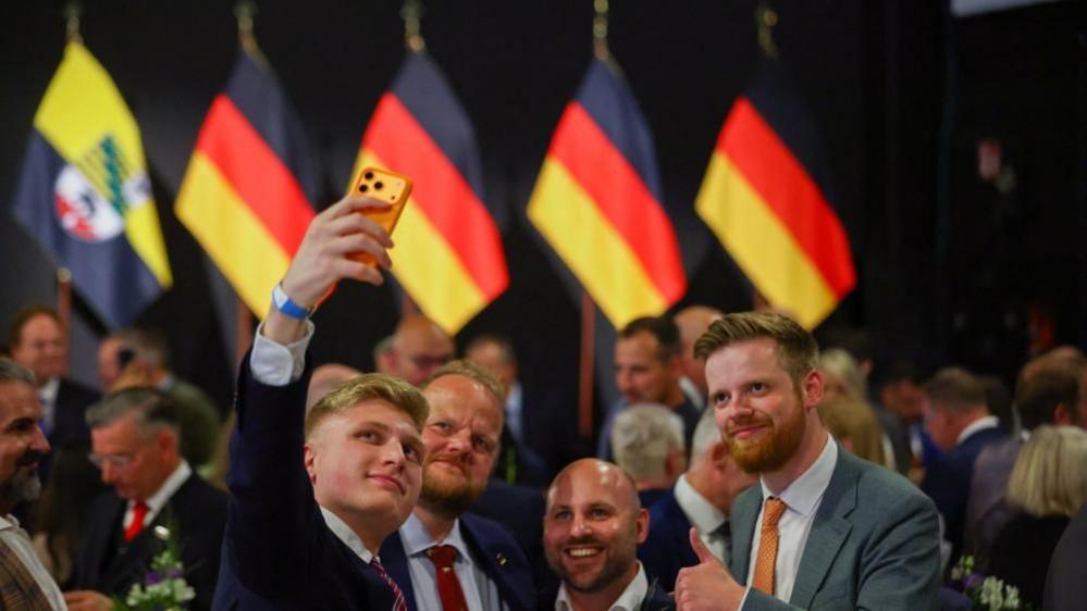 Far-right Alternative for Germany (AfD) supporters pose for a selfie at a venue being used by the AfD, on the day of the Saxony-Anhalt state elections, in Magdeburg, Germany, with German flags flying behind them