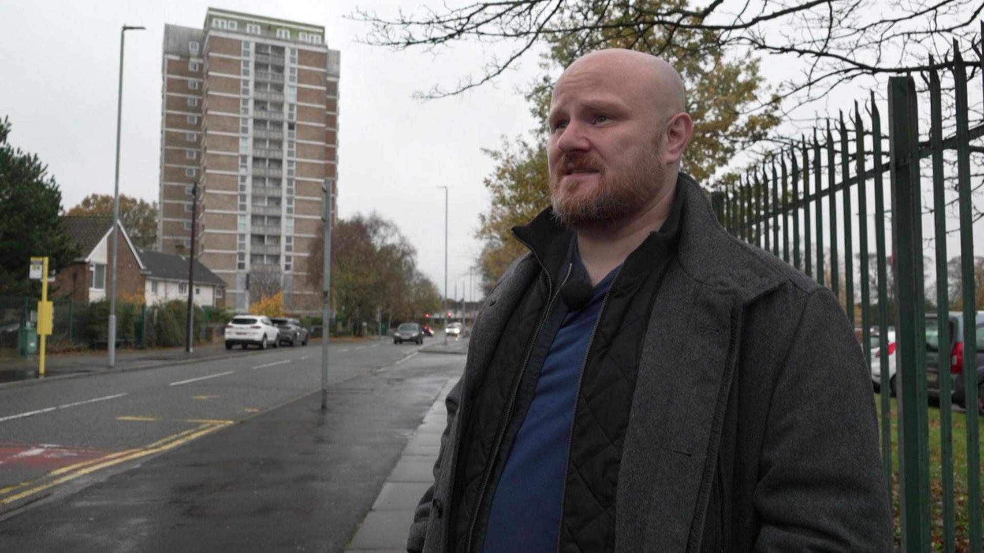 Michael Jones, who is bald and has a short ginger beard, is standing outside Willow Rise in Kirkby. The building is a 1960s tower block. Mr Jones is looking at a reporter who is off camera.