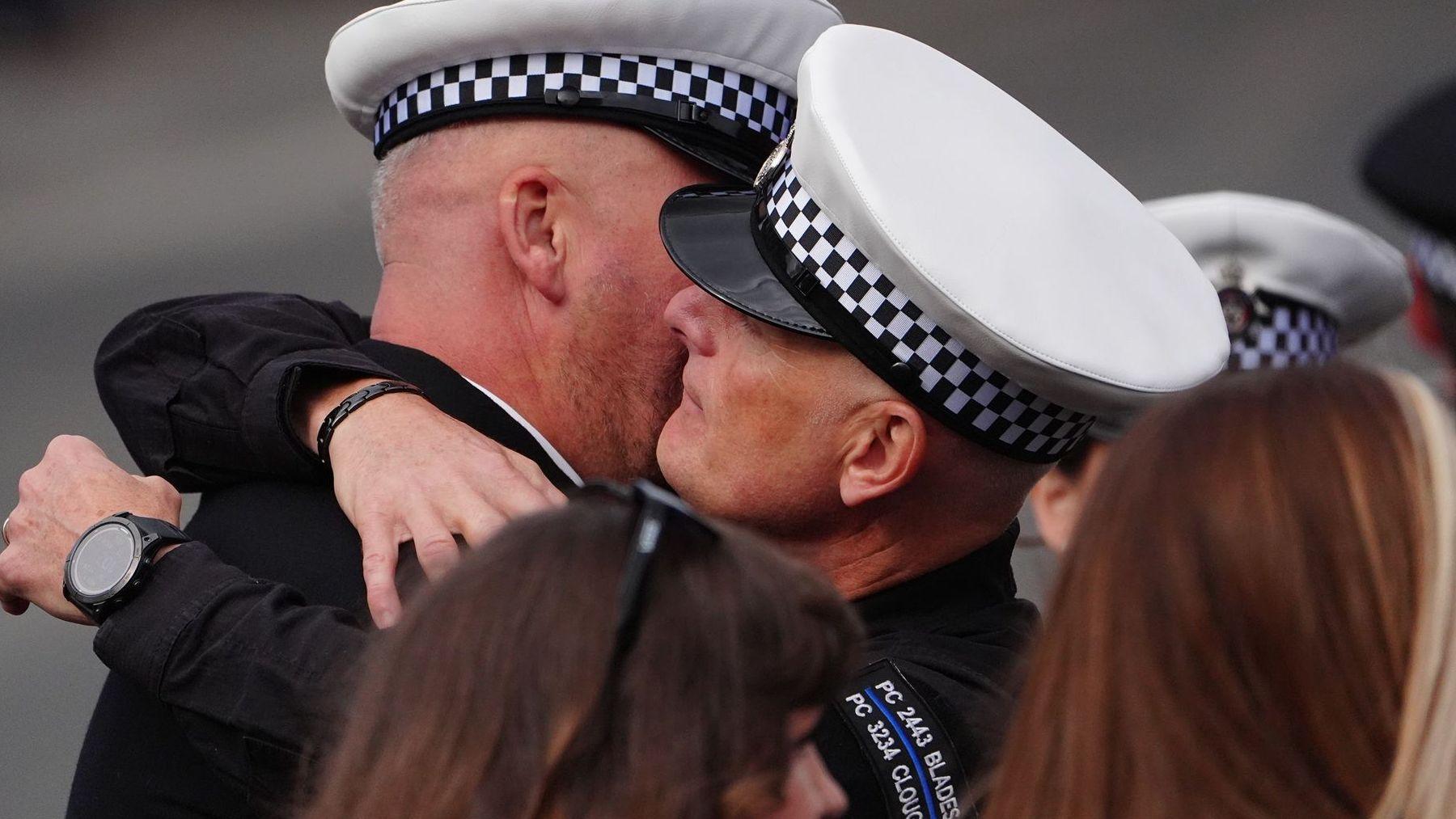 Two male police officers embracing. Both are wearing black uniforms with white capped police hats. Bother men are bald and the man on the left's face is hidden by the man on the right. The man on the right is wearing one of the PC Blades and PC Clough patches. The heads of two women are in front of them with brown hair and are partially cut off.