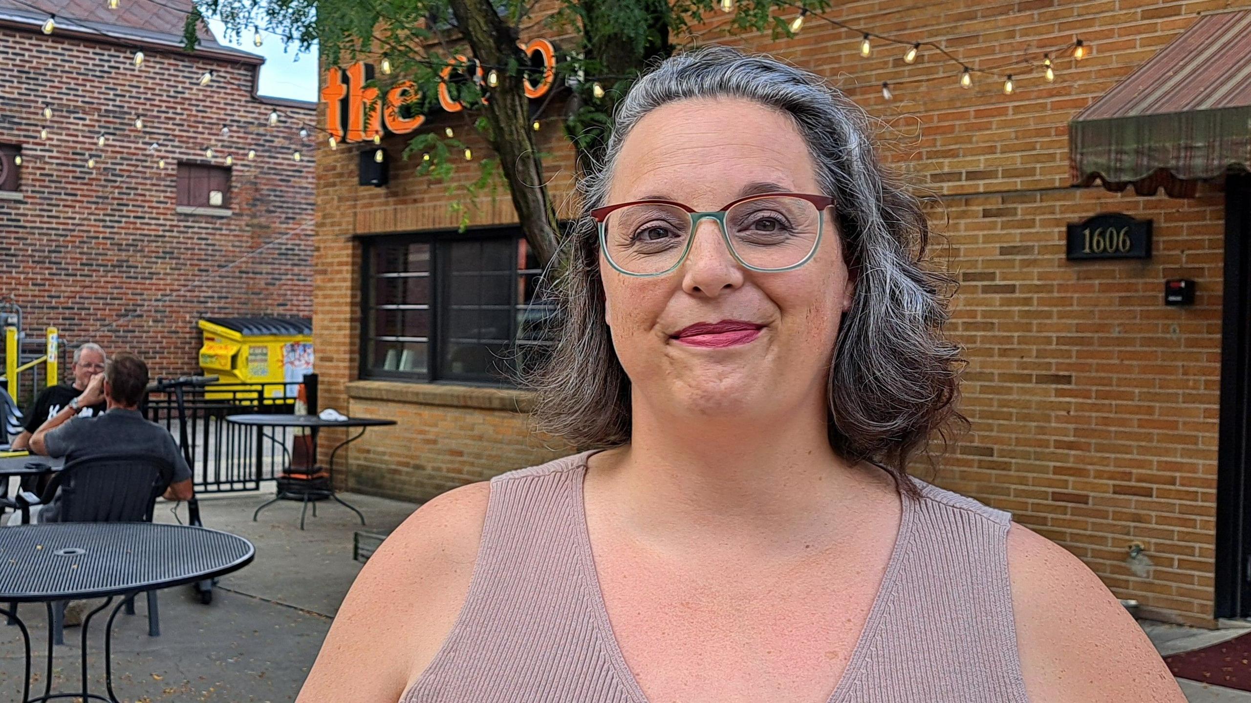 Brianna Beyrouti, wearing glasses and smiling, standing in front of what appears to be a cafe or restaurant, with two men sitting at outdoor tables behind her
