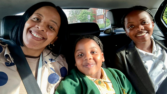 A smiling family sitting together in the back seat of a car. To the left is the mum, wearing a blue polka dot jacket. In the middle is a young girl in a green and yellow school uniform. On the right is an older girl in a white shirt and grey blazer.