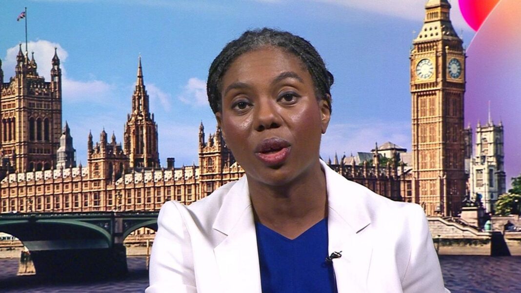 A head and shoulders shot of Badenoch in a BBC studio. She is wearing a white jacket over a royal blue top and has her braided hair tied back in a low ponytail.
