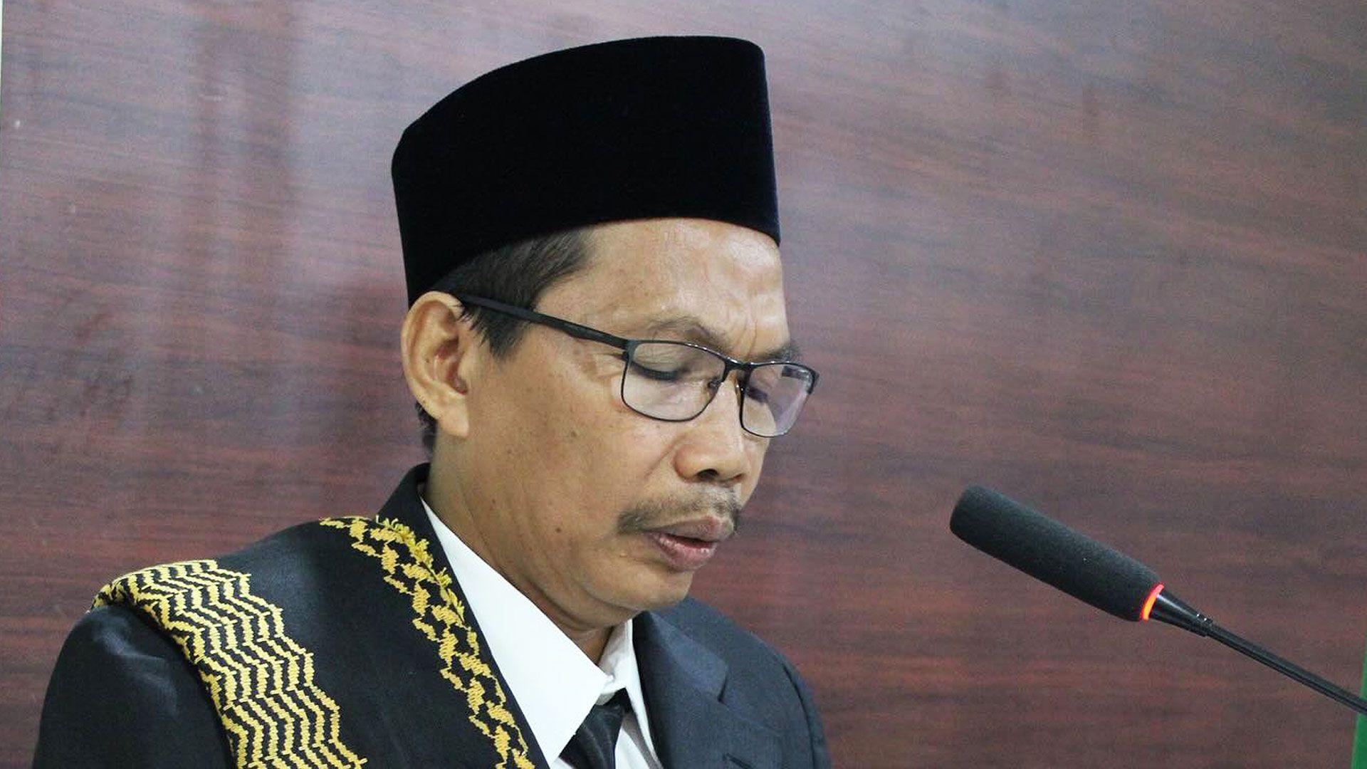 Abah wears a black kopi on his head, and a black suit with black tie and white shirts, with a black and yellow sash draped over one shoulder. He is speaking at a podium, a microphone in front of him.