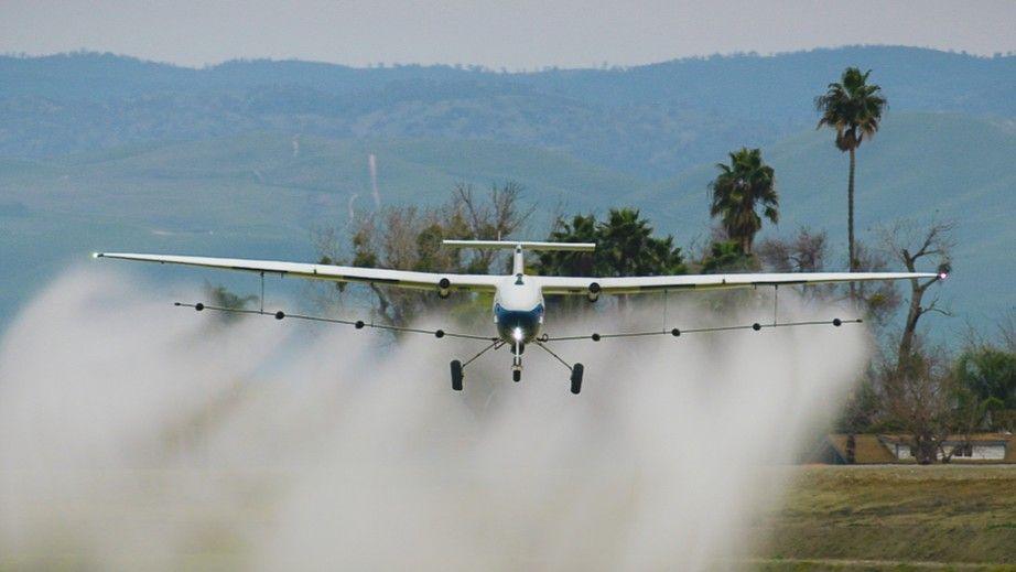 A Pyka crop-duster flies low over a field spraying liquid.