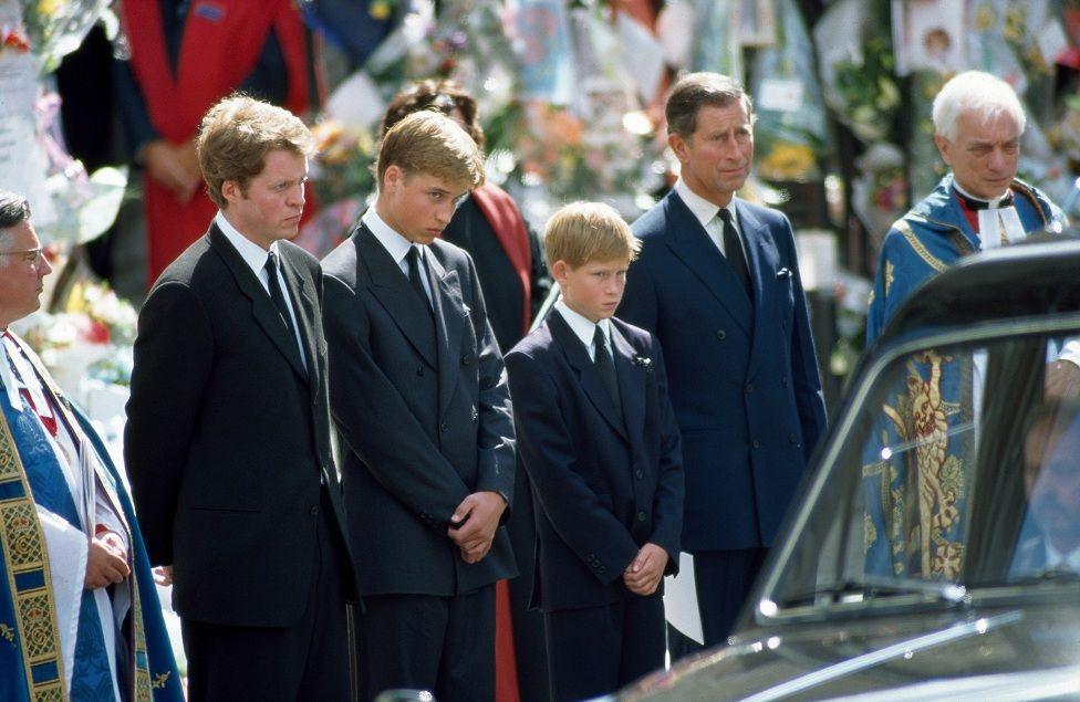 Earl Spencer with Princes William and Harry, and Prince Charles at the funeral of Diana, Princess of Wales, standing alongside members of the clergy to the side of the funeral procession, with floral tributes faintly visible in the background