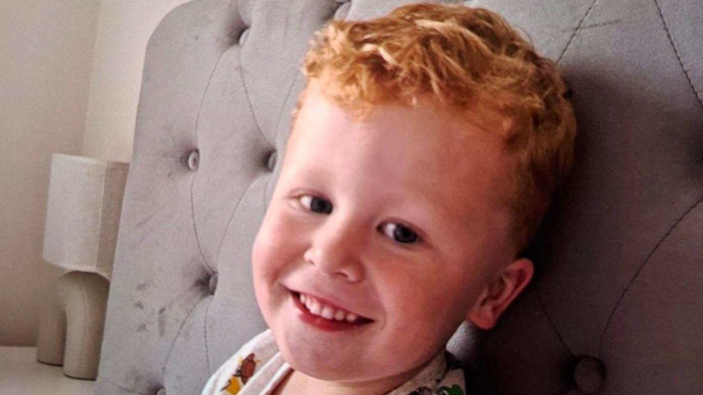 Noah, who has ginger hair, is pictured smiling on a bed.