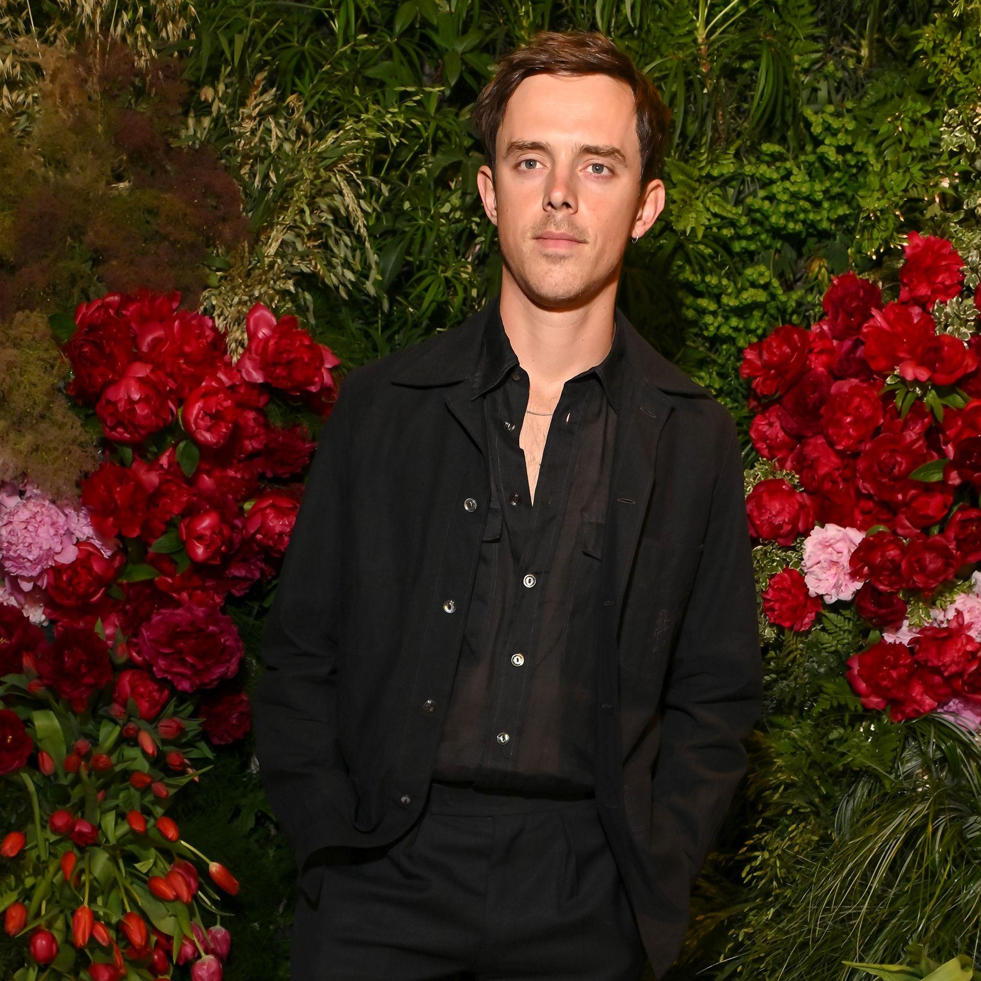 Jack Barton looks at the camera while wearing a black shirt against a shadowed flowers background, in London in May.