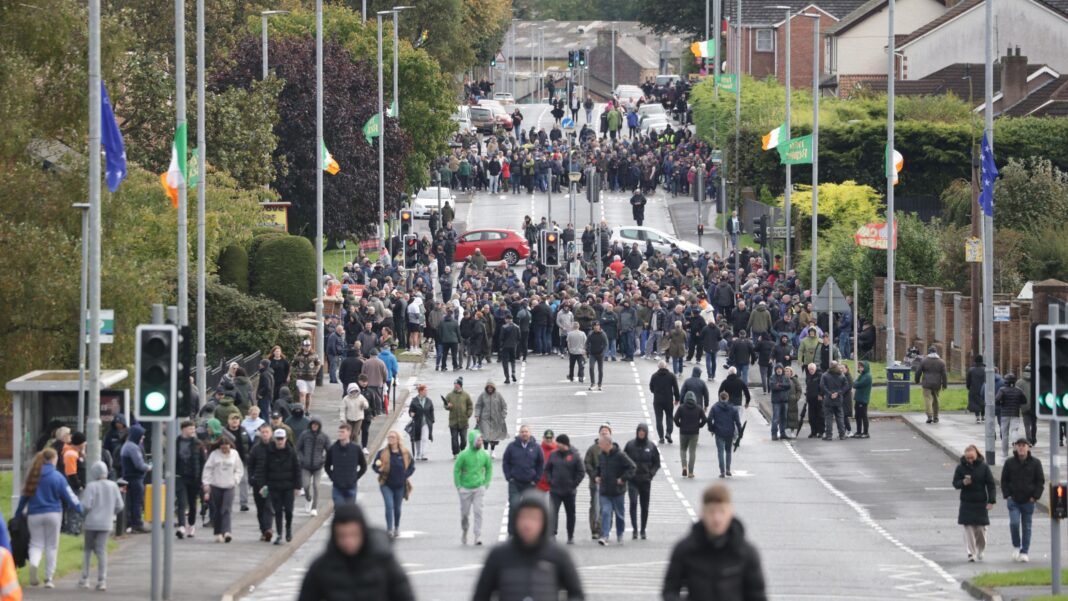A wide angle shot showing large crowds on the Garvaghy Road, split out onto two distinct groups, spread across the length of the road, blocking access. There are also some vehicles parked across the road, again blocking it. The street lamps lining the road, which features greenery and red bricked homes, are adorned with Irish green-white-orange tricolor and blue starry plough flags.