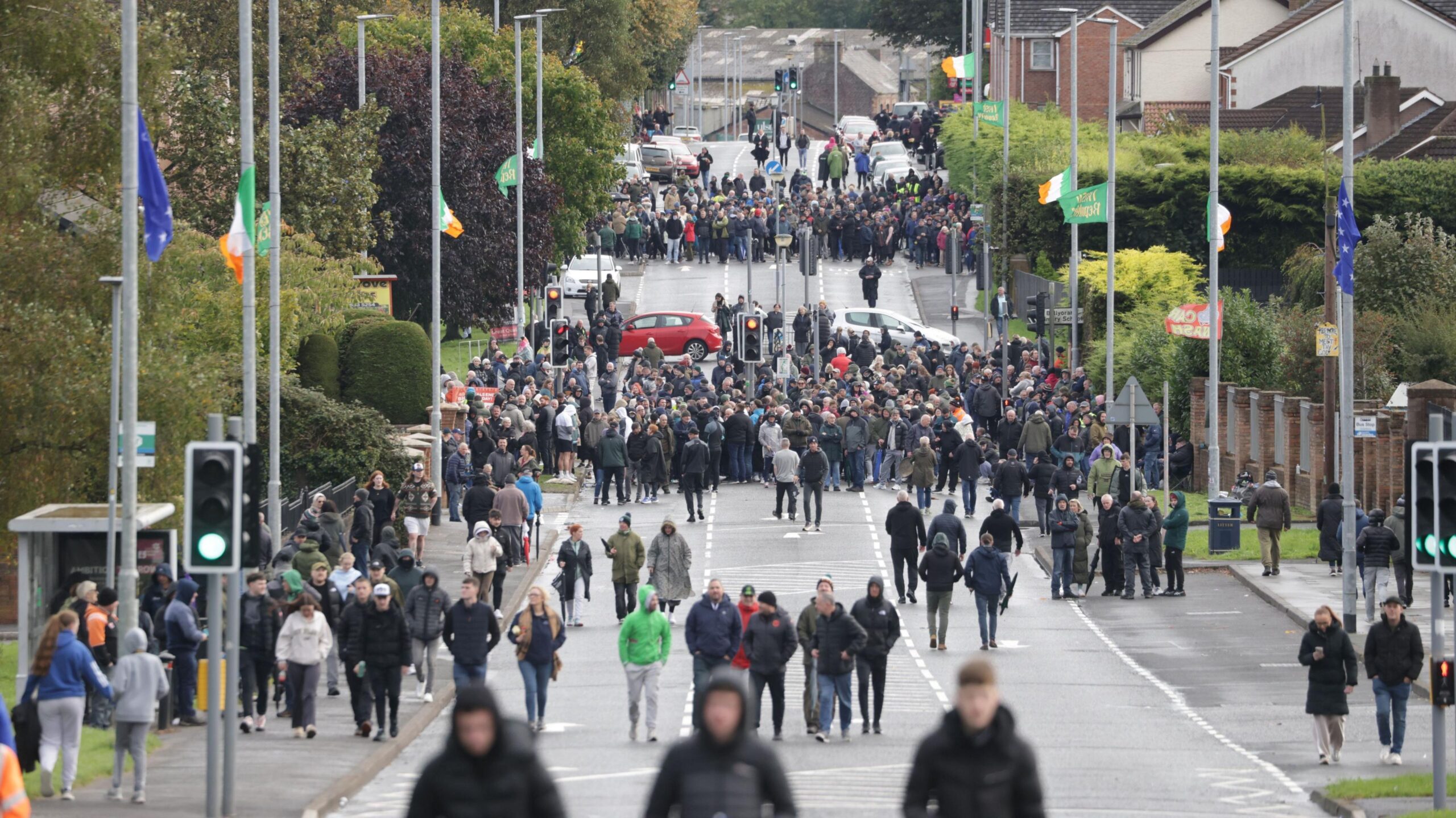 A wide angle shot showing large crowds on the Garvaghy Road, split out onto two distinct groups, spread across the length of the road, blocking access. There are also some vehicles parked across the road, again blocking it. The street lamps lining the road, which features greenery and red bricked homes, are adorned with Irish green-white-orange tricolor and blue starry plough flags.