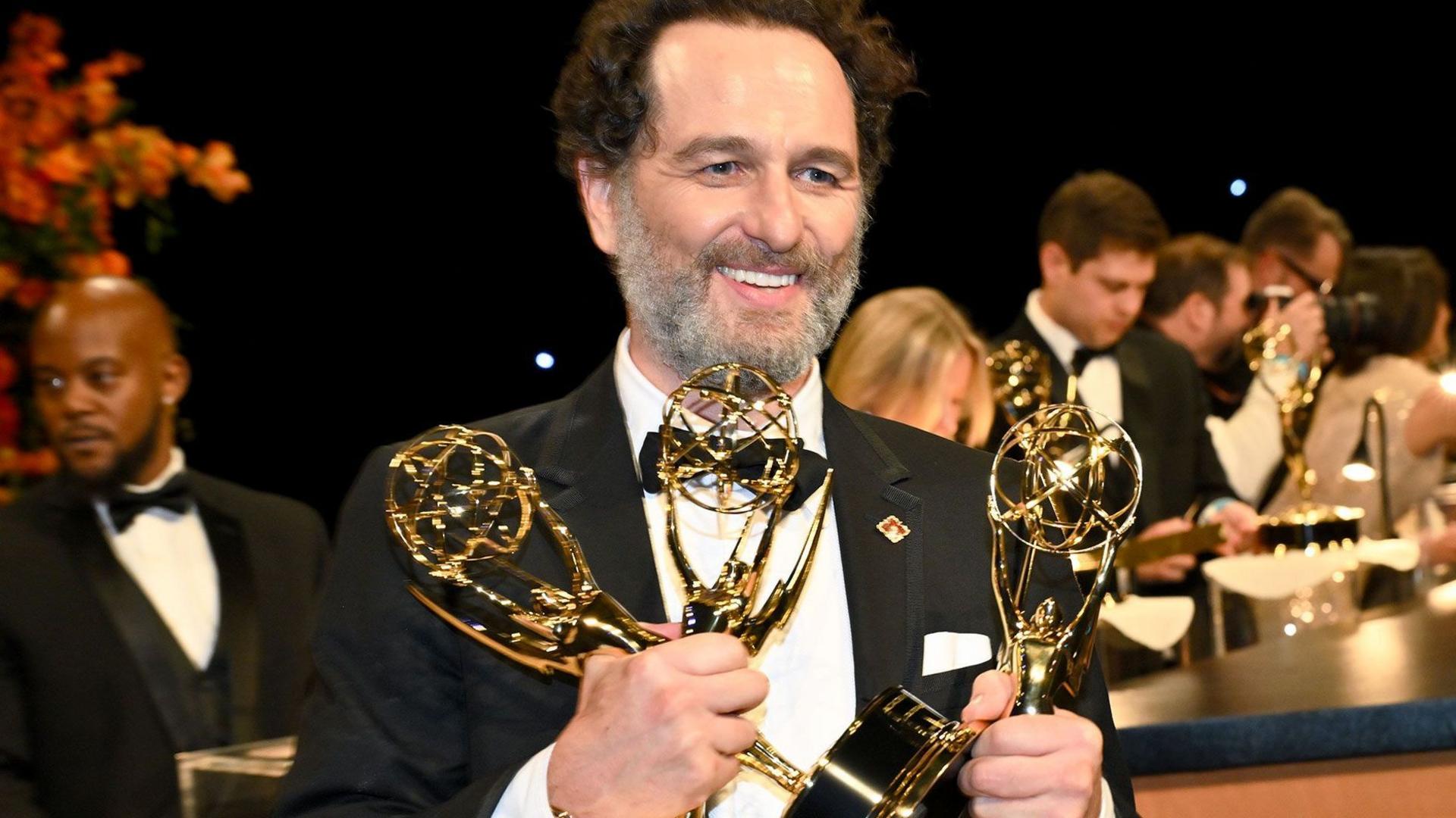 Matthew Rhys is photographed attending the 78th Emmy Awards. He is wearing a black suit jacket, black waist coat and black bow tie. He has dark hair and a grey beard. He is smiling whilst holding three Emmy awards. He is looking away from the camera.