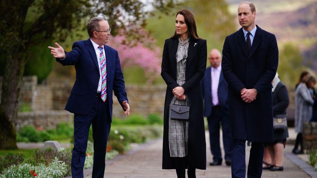 A man in a navy suit and colourful tie raises an arm to guide the Prince and Princess of Wales through the memorial garden