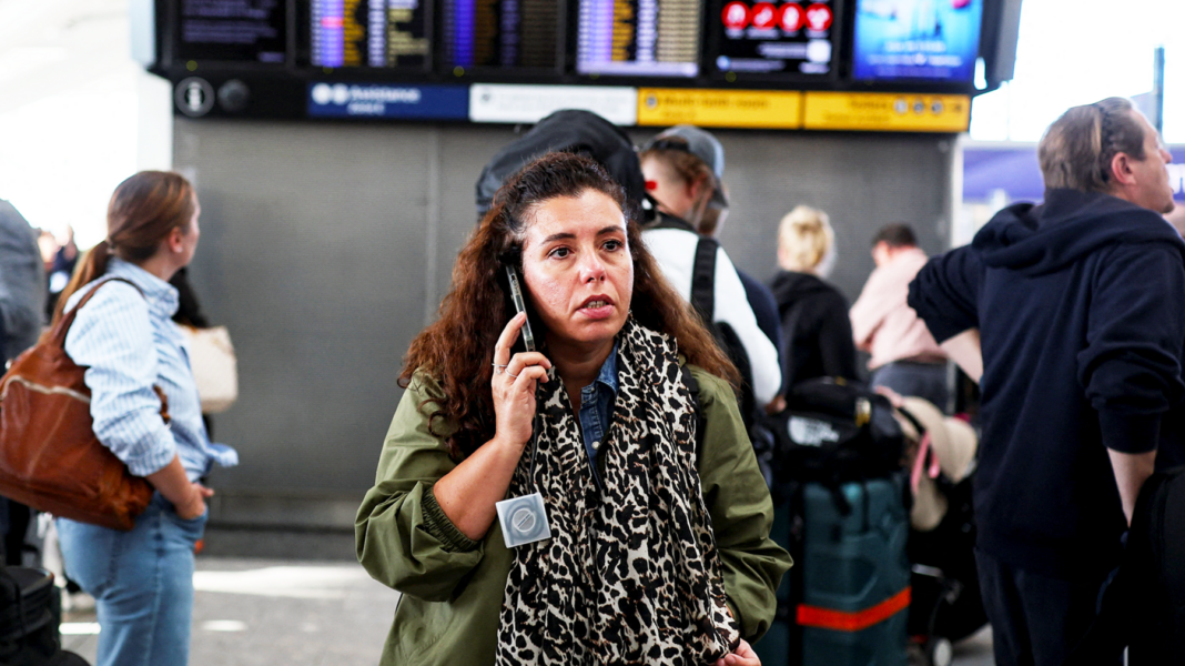 A woman wearing a green coat and leopard print scarf talks on the phone inside an airport with flight information on TV screens behind her