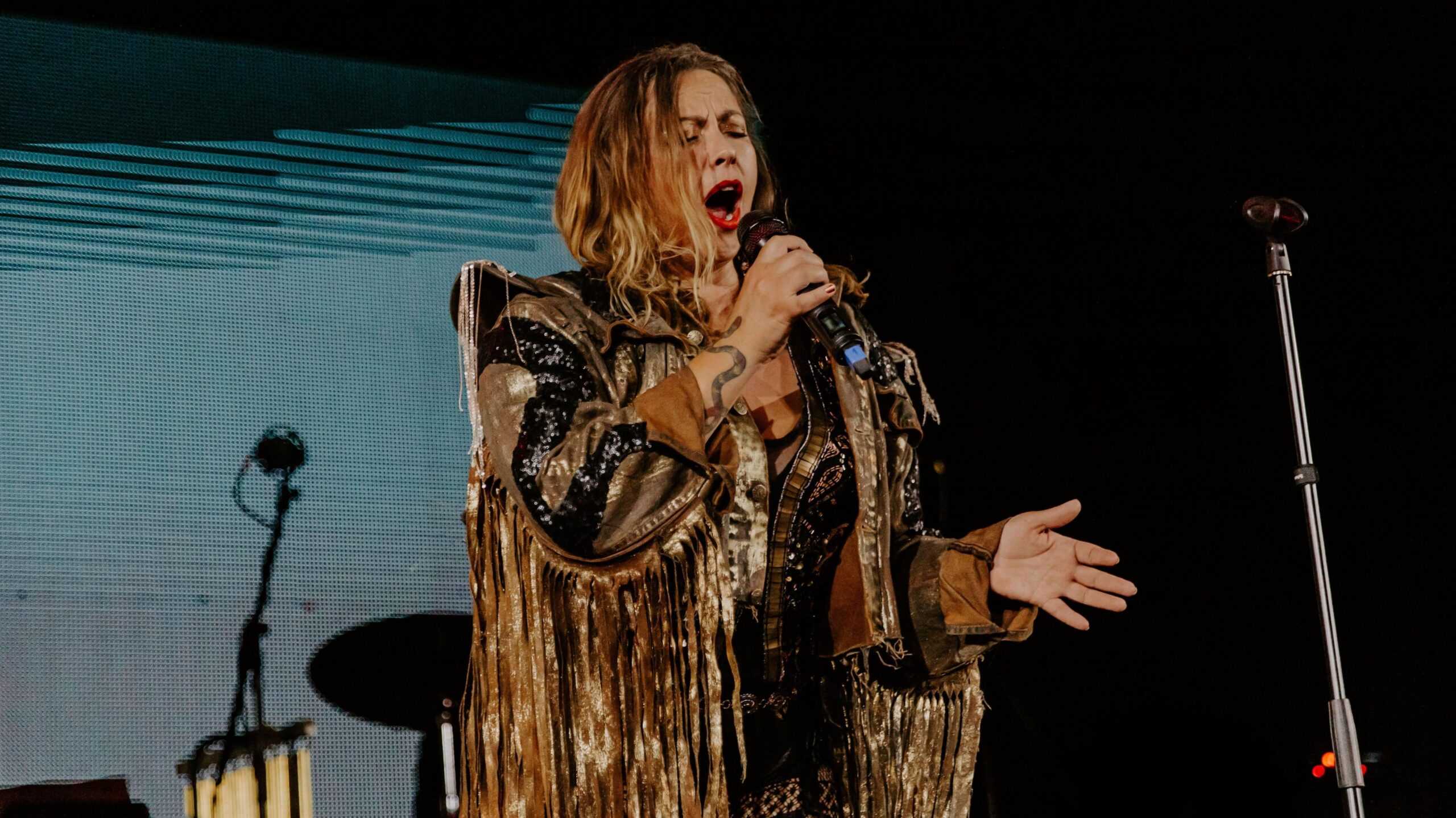 Charlotte Church singing on stage, she is wearing a gold and black sequin jacket with gold tassels on. She has long blonde hair.
