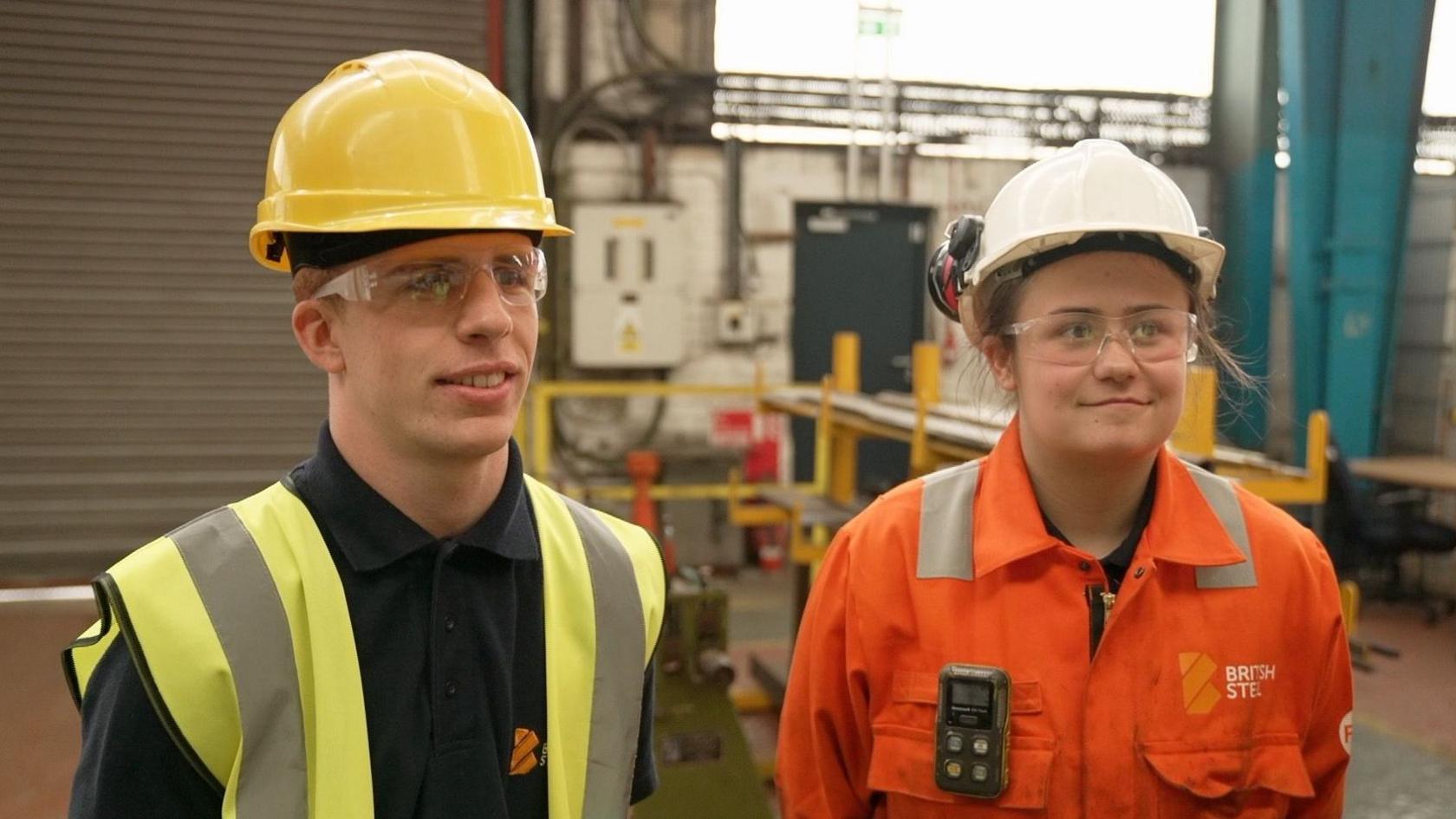Two apprentices standing inside a warehouse, smiling at someone to the right of the camera. The man on the left is wearing a yellow hard hat, a black polo shirt and a yellow hi-vis vest. The woman on the right is wearing a white hard hat and an orange hi-vis jacket.