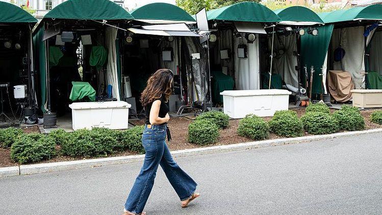 A woman walking past a row of tented booths, with cameras and media equipment inside, set up on the White House grounds