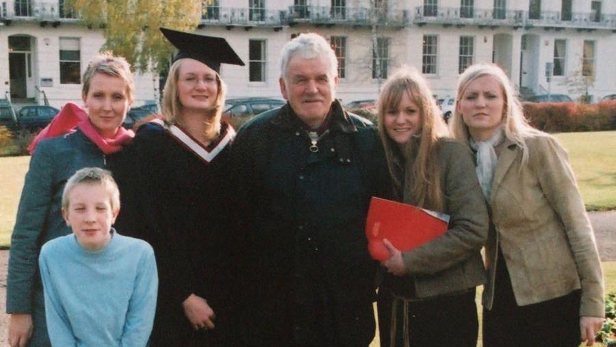 An old photo of a family standing together. Michael Gwilliam is in the middle of his four daughters, with one of them wearing a graduation cap and gown. A young boy crouches down in front of them.