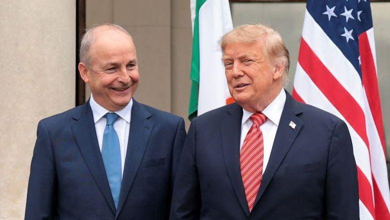 Micheál Martin to the left with blue suit and blue tie. Donald Trump to his right with dark suit and red tie. The Irish and American flag are behind them.