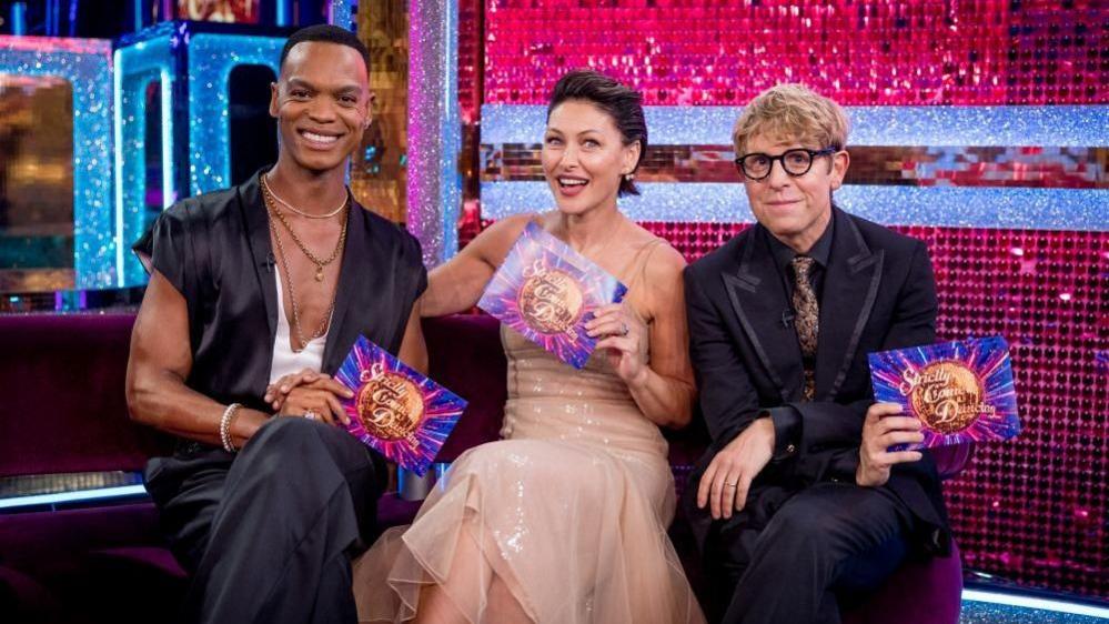 Left to right: Johannes Radebe, Emma Willis and Josh Widdicombe sit in a line on a sofa.