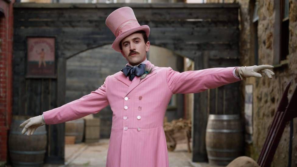 Callum Scott Howells dressed in one of Henry Paget's outfits. It is a pink buttoned up suit jacket paired with a large navy blue bow tie. He is also wearing a pink top hat and has white gloves on his outstretched hands.