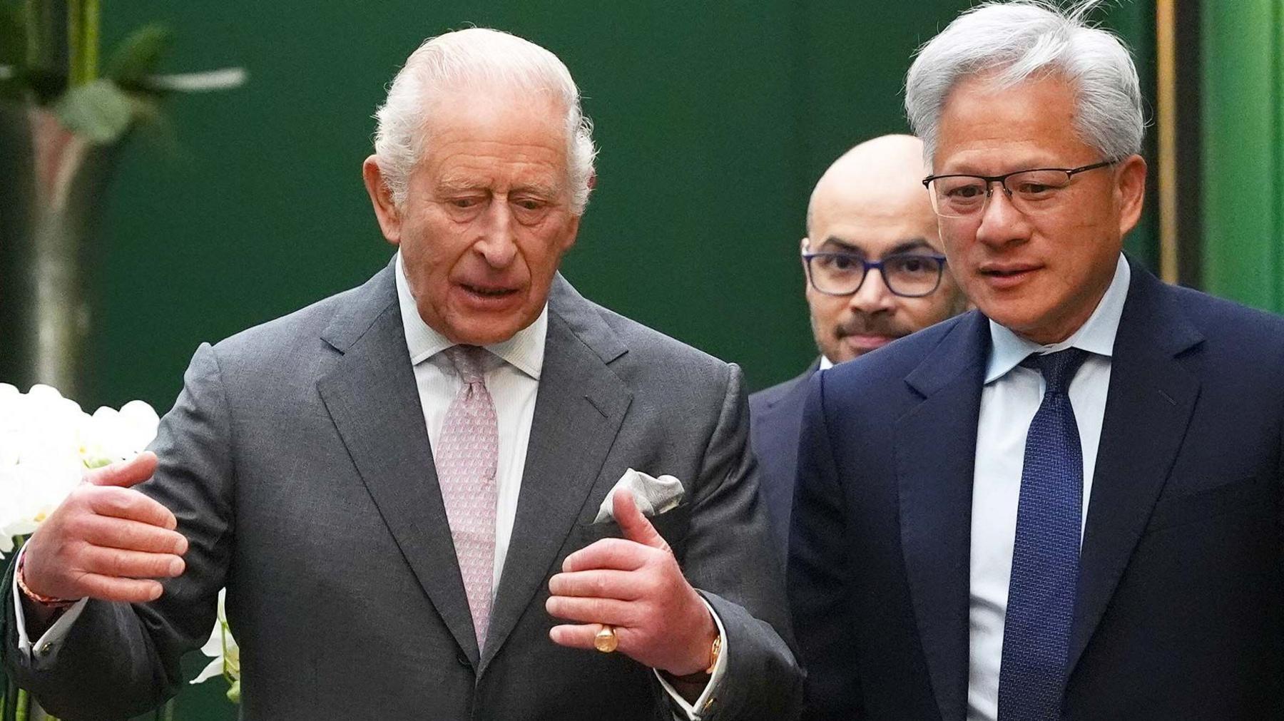 King Charles wearing a grey suit jacket and white shirt and pink tie, talking to Jensen Huang, the CEO of Nvidia, who is wearing a dark suit, white shirt and blue tie. Former Google DeepMind boss Sir Demis Hassabis is walking behind them.