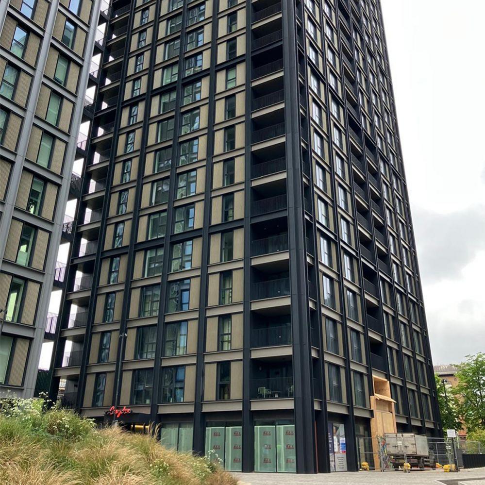 A tall modern high‑rise with a grid of dark metal framing and large glass windows. Several balconies punctuate the facade. At ground level, glass doors and windows—some covered for ongoing work—sit above landscaped grass and plants, with construction materials nearby.