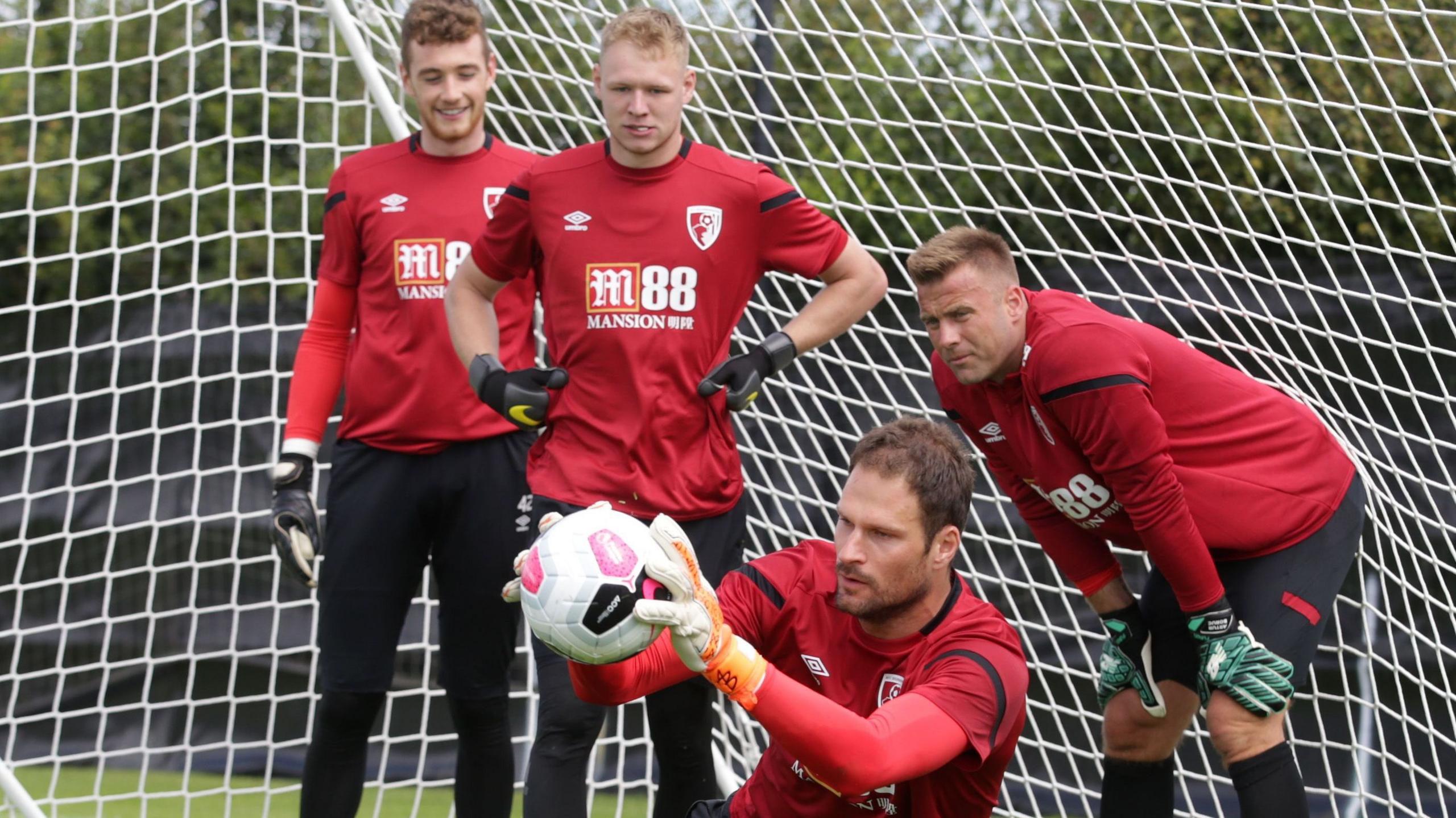 Pictured in 2019, Aaron Ramsdale, wearing a red Bournemouth training top, watches on in training as Asmir Begovic, also wearing a red Bournemouth training top and gloves catches a ball while making a save
