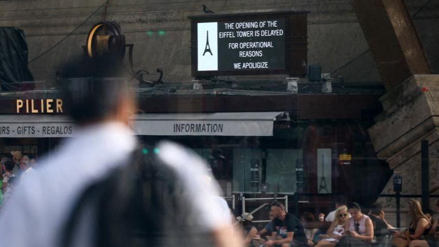 A man wearing a backpack is out of focus in the foreground of the picture, which shows the entrance to the Eiffel Tower and a sign showing that it is closed.