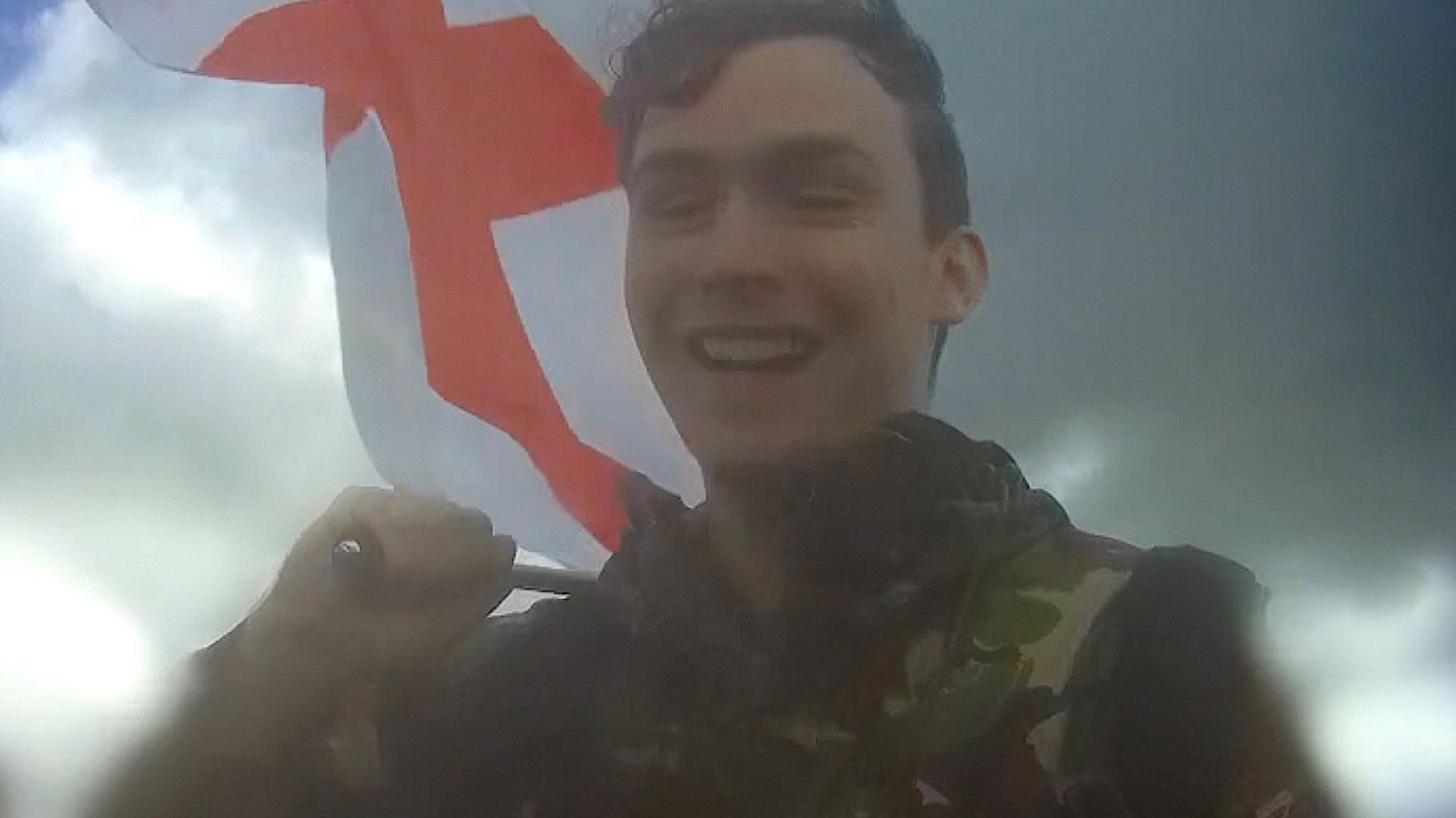 Webster, smiling, wearing a camouflage top and holding an England flag over one shoulder