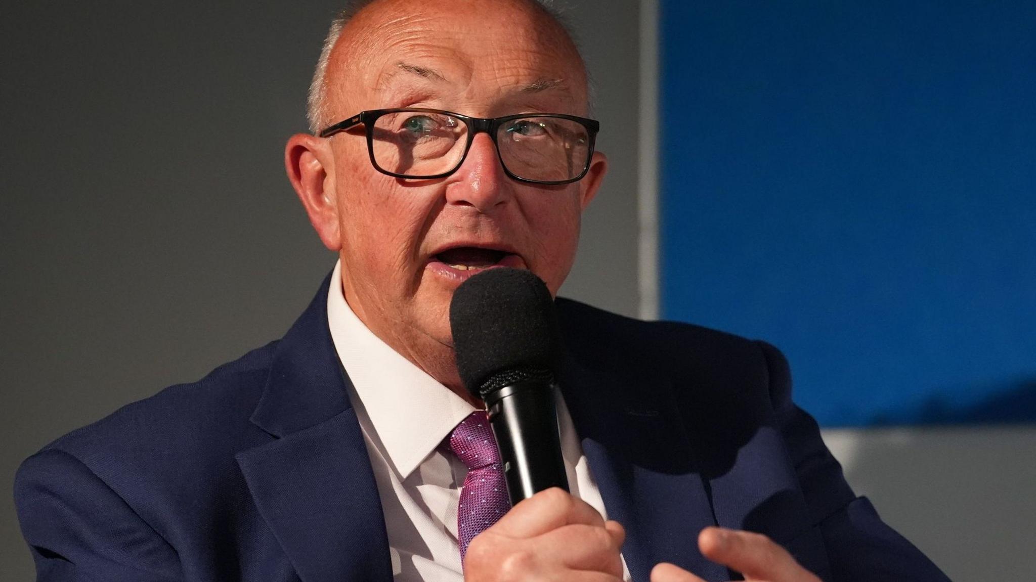 Colin Sutton is wearing black rimmed glasses and a navy suit, white shirt and purple patterned tie. He is holding a microphone close to his mouth and looks to be in a corporate environment.