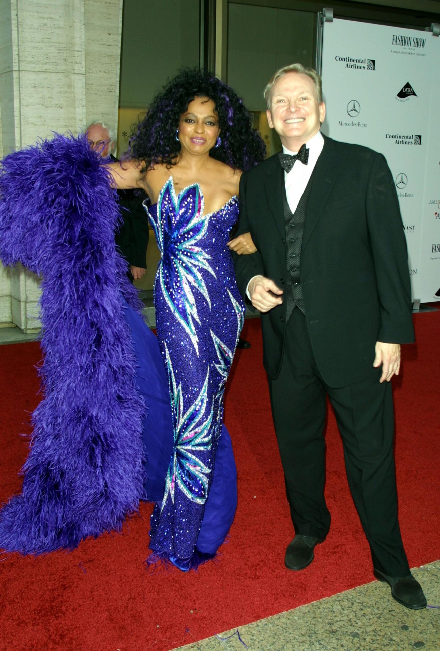 Diana Ross on the red carpet at the 20th CFDA American Fashion Awards in 2001, arm-in-arm with Bob Mackie. She wears a long blue dress and carries a sapphire feather boa.