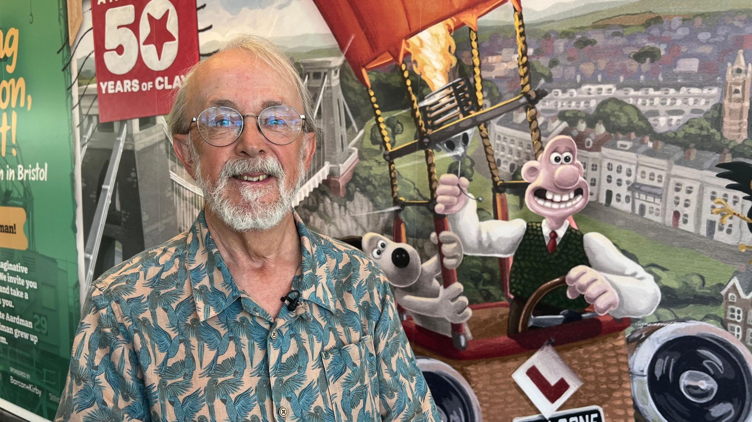 Sir Peter Lord is wearing a shirt with blue parrots on it. He has a white bear and hair and is wearing round glasses. He is standing in front of a poster where Wallace and Gromit are pictured riding in a hot air balloon.