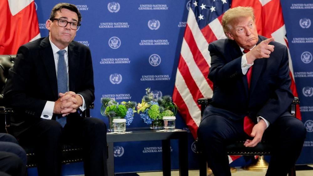 Andy Burnham is sat on the left and Donald Trump on the right. Both men are wearing dark coloured suits and white shirts. Burnham has a blue tie and is wearing glasses, Trump has a red tie. Trump is pointing in the direction of the journalists, who are not pictured. There is a Union flag and US flag behind each man. The background is blue and has "United Nations General Assembly" written on it several times. There is a table between the two men. There are floral decorations and glasses of water on the table.